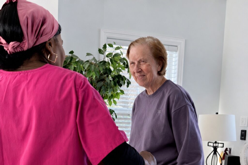 Caregiver in pink scrubs warmly greeting a resident in a bright room