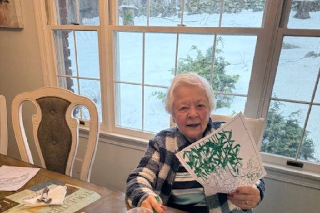 Resident seated at a dining table beside a snowy window, smiling and holding up a handmade card