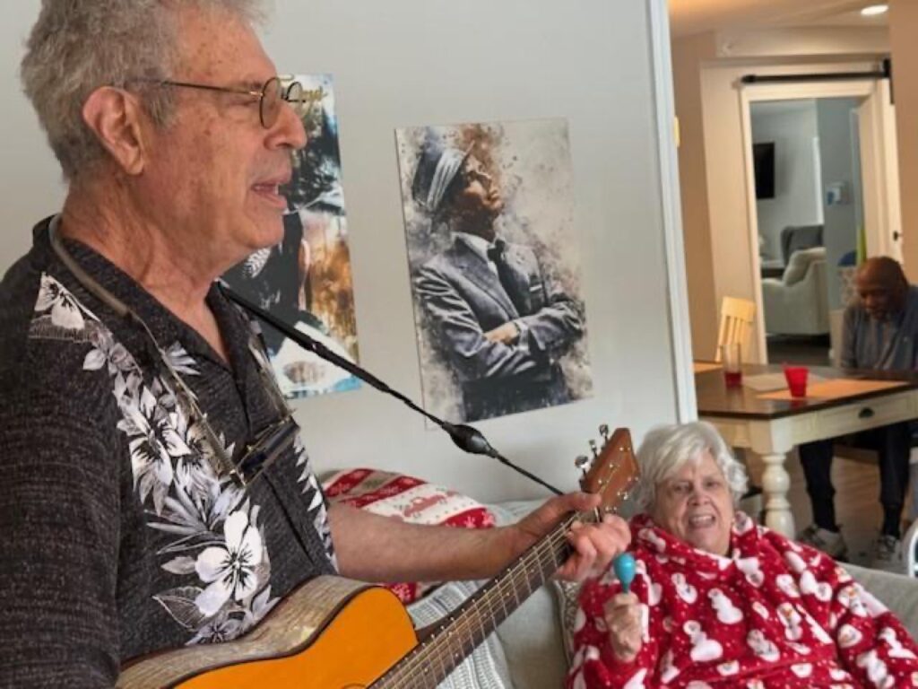 Musician playing acoustic guitar and singing for a resident relaxing in a nearby chair