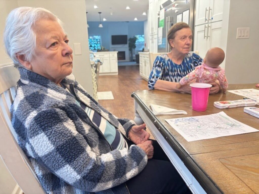 Two residents at the dining table with coloring pages, one holding a baby doll